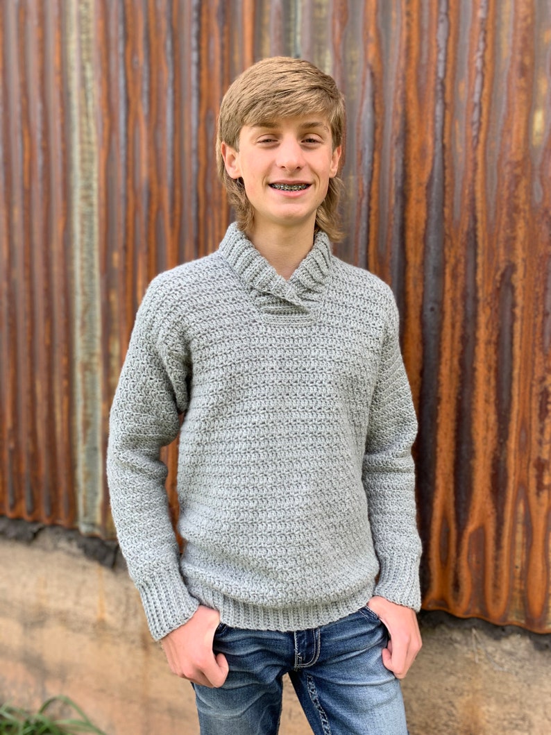 Pu&ograve; includere: Un maglione all'uncinetto grigio chiaro con collo a scialle. Il maglione ha maniche lunghe e orlo e polsini a coste. La persona indossa jeans blu. Lo sfondo &egrave; un muro di metallo arrugginito.