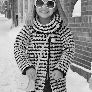 May include: A young girl wearing a black and white houndstooth coat with a white headband and sunglasses. She is holding a white purse with a black strap.