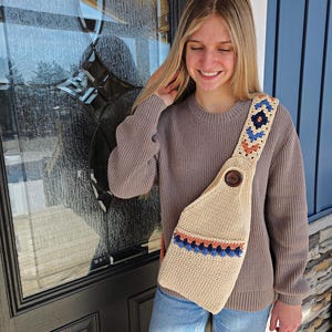 May include: Beige crochet shoulder bag with a decorative strap featuring blue, orange, and cream-colored geometric patterns. The bag has a large wooden button closure and a matching crochet strap.