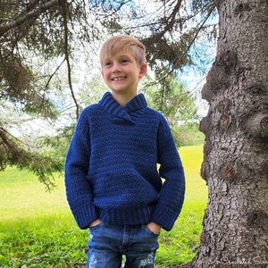 May include: A young boy wearing a blue crocheted sweater with a collared neckline, standing in a grassy field with a tree behind him.