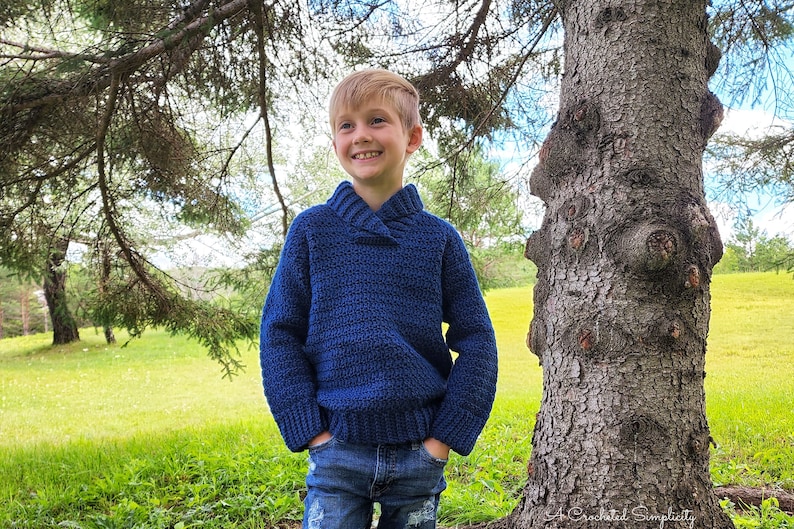 May include: A young boy wearing a blue crocheted sweater with a collared neckline, standing in a grassy field with a tree behind him.