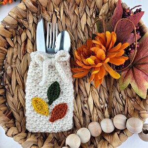 May include: A cream-coloured, knitted cutlery holder with three embroidered leaves in yellow, green, and orange. The holder contains a knife, fork, and spoon. The setting includes an orange flower and decorative autumn elements.