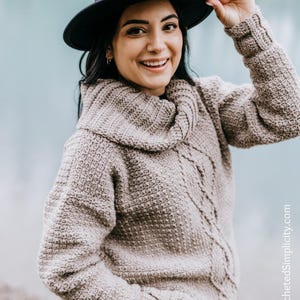 Op de afbeelding: Een vrouw draagt een beige gebreide trui met een col en kabelbreisteek. Ze draagt een zwarte fedora-hoed en een blauwe spijkerbroek. De trui is grof gebreid en heeft een losse pasvorm.