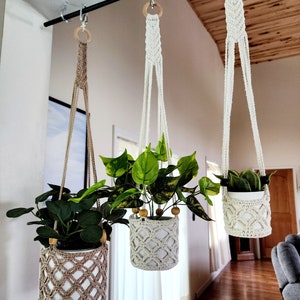 May include: Three macrame plant hangers with wooden rings hanging from a black metal rod. Each hanger holds a potted plant with green leaves. The hangers are made with a white and beige cord.