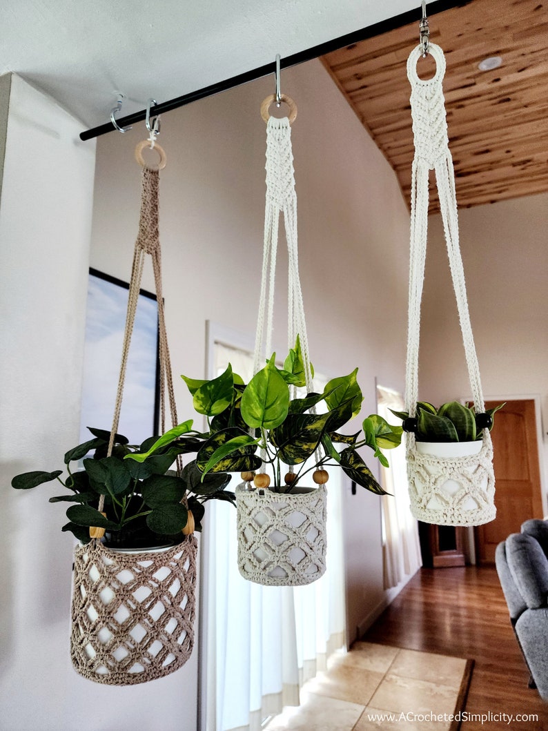 May include: Three macrame plant hangers with wooden rings hanging from a black metal rod. Each hanger holds a potted plant with green leaves. The hangers are made with a white and beige cord.