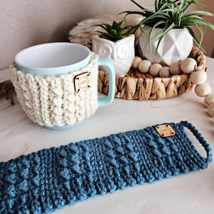 May include: A light blue ceramic mug with a white crocheted cozy and a wooden tag that says "MUGS". A blue crocheted mug cozy is also shown.
