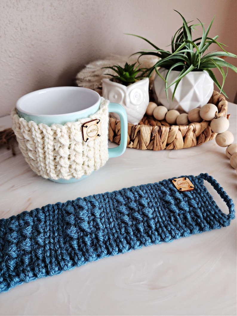 May include: A light blue ceramic mug with a white crocheted cozy and a wooden tag that says "MUGS". A blue crocheted mug cozy is also shown.