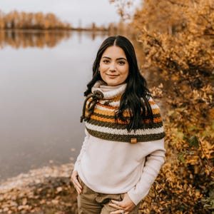 May include: A woman wearing a white sweater and brown pants stands in front of a lake with fall foliage. She is wearing a crocheted cowl with green, white, orange, and brown stripes.