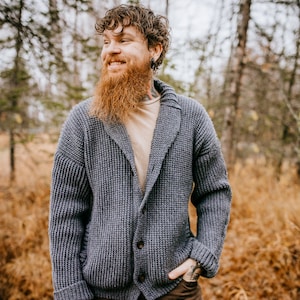 May include: A man wearing a gray knitted cardigan with a shawl collar and brown pants. He is standing in a forest with fall foliage.