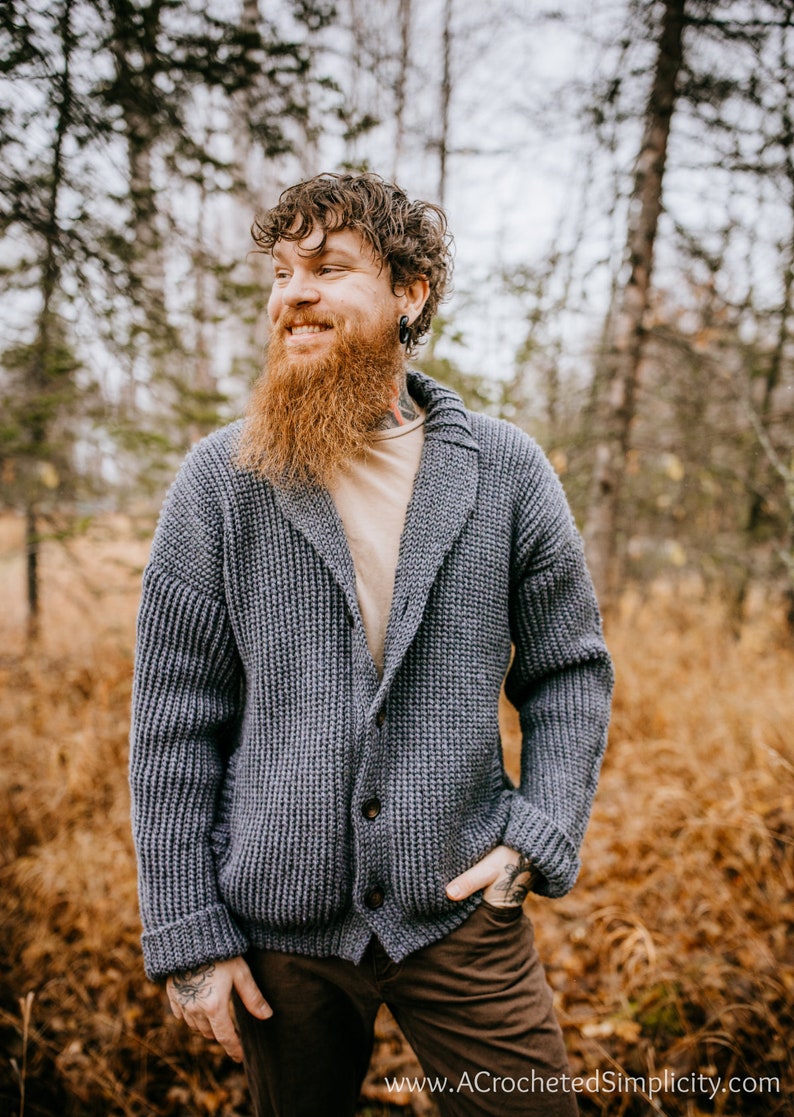 May include: A man wearing a gray knitted cardigan with a shawl collar and brown pants. He is standing in a forest with fall foliage.