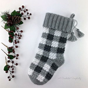 May include: A gray and white crocheted Christmas stocking with a black and white buffalo plaid pattern. The stocking has a gray crocheted cuff and a gray tassel.