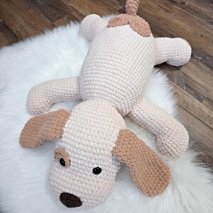 Puede incluir: Un juguete de peluche de perro hecho a mano con ganchillo en crema y marr&oacute;n claro. El perro tiene orejas ca&iacute;das, una nariz marr&oacute;n y ojos negros. El juguete est&aacute; sobre una alfombra blanca y esponjosa.