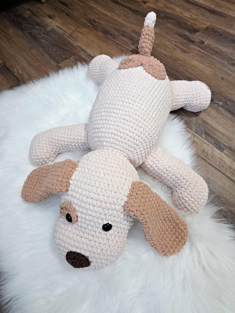 Puede incluir: Un juguete de peluche de perro hecho a mano con ganchillo en crema y marr&oacute;n claro. El perro tiene orejas ca&iacute;das, una nariz marr&oacute;n y ojos negros. El juguete est&aacute; sobre una alfombra blanca y esponjosa.