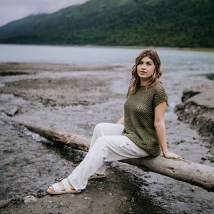 May include: A woman wearing a green crochet top and white pants sits on a log by a river. The river is surrounded by green hills and a cloudy sky.