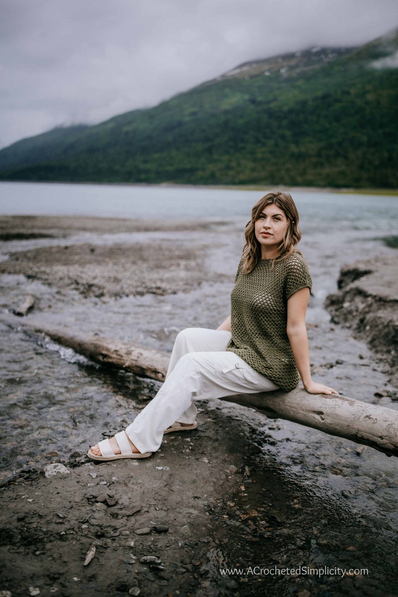 May include: A woman wearing a green crochet top and white pants sits on a log by a river. The river is surrounded by green hills and a cloudy sky.