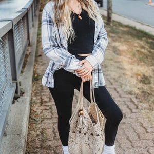 May include: A beige crochet tote bag with a tassel and wooden bead accents. The bag is held by a person wearing black leggings, a black top, a blue and white plaid shirt, and gray suede shoes.