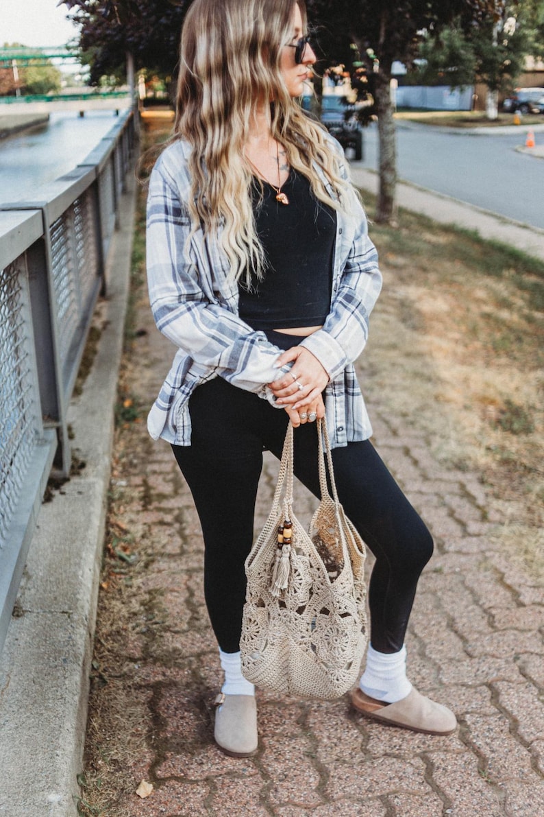 May include: A beige crochet tote bag with a tassel and wooden bead accents. The bag is held by a person wearing black leggings, a black top, a blue and white plaid shirt, and gray suede shoes.