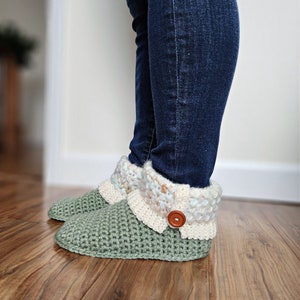 May include: Green crocheted slippers with a white and blue crocheted cuff and a brown button. The slippers are worn by a person wearing blue jeans.