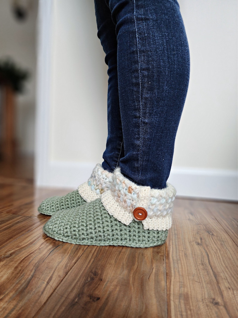 May include: Green crocheted slippers with a white and blue crocheted cuff and a brown button. The slippers are worn by a person wearing blue jeans.