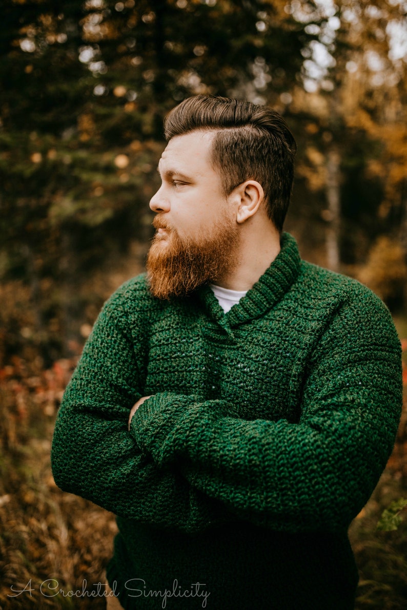 Pu&ograve; includere: Un maglione verde lavorato a mano all'uncinetto con collo a scialle. Il maglione &egrave; di un colore verde foresta intenso e presenta un motivo a punto strutturato. L'immagine include il testo "A Crocheted Simplicity".
