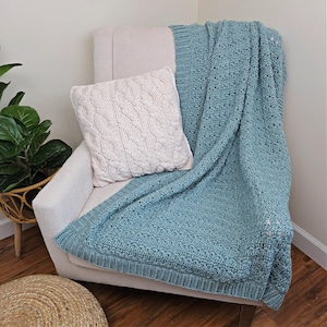 May include: A light blue crocheted blanket draped over a white armchair, paired with a cream-colored cable-knit pillow. The blanket has a textured stitch pattern and a ribbed border. The pillow features a detailed cable design, creating a cozy, inviting aesthetic.