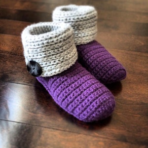 May include: A pair of handmade crochet slippers. The slippers have a purple foot and a gray cuff. A black button is attached to the cuff. The slippers are made of a soft yarn and are designed for indoor use.