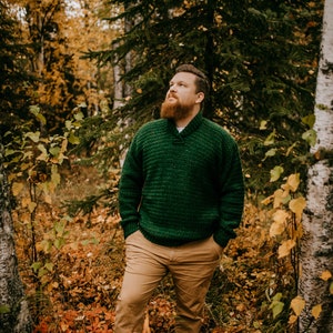 Pu&ograve; includere: Una persona indossa un maglione verde lavorato a maglia con collo a scialle e pantaloni color cachi in una foresta. Il maglione ha un motivo strutturato. Il suolo della foresta &egrave; coperto di foglie cadute. Il testo "A Crocheted Simplicity" &egrave; in basso.