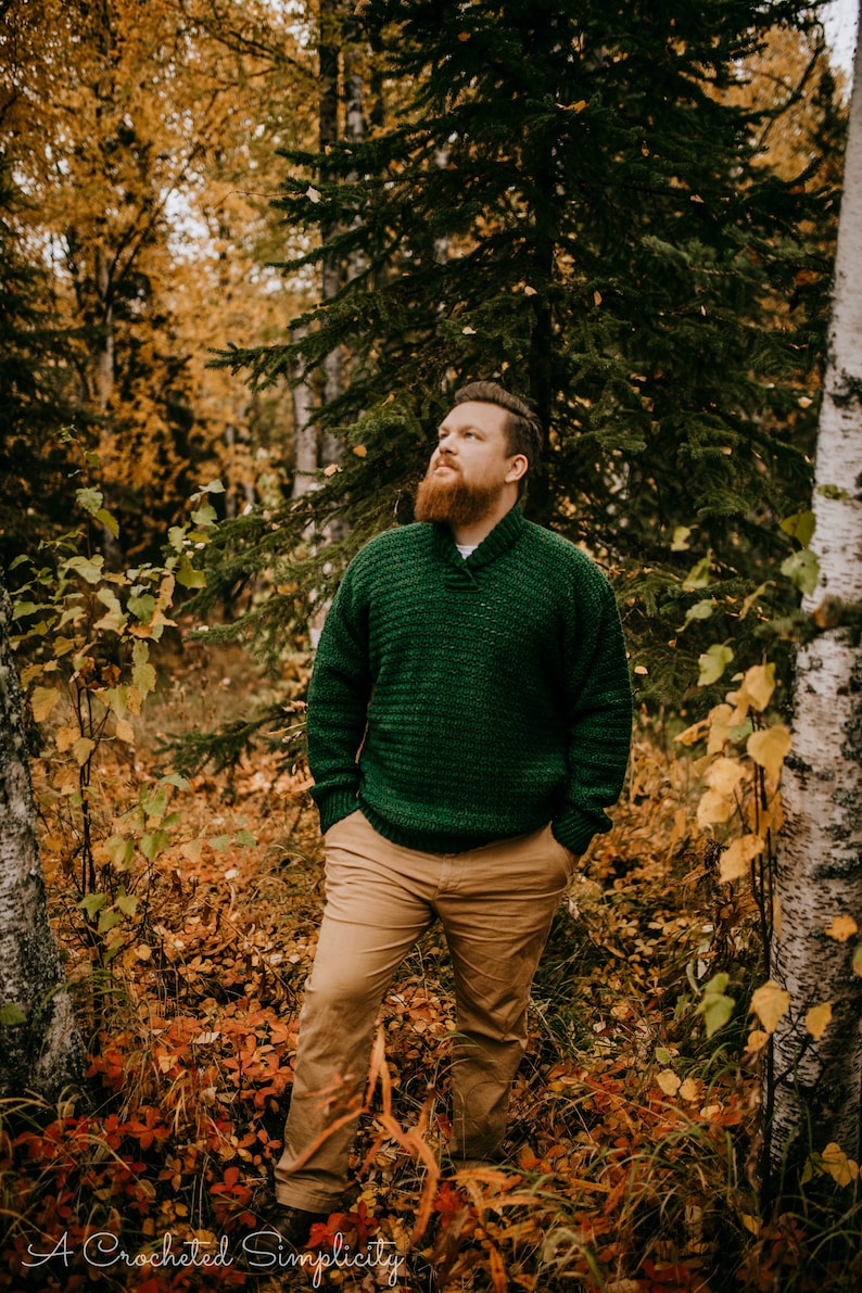 Pu&ograve; includere: Una persona indossa un maglione verde lavorato a maglia con collo a scialle e pantaloni color cachi in una foresta. Il maglione ha un motivo strutturato. Il suolo della foresta &egrave; coperto di foglie cadute. Il testo "A Crocheted Simplicity" &egrave; in basso.