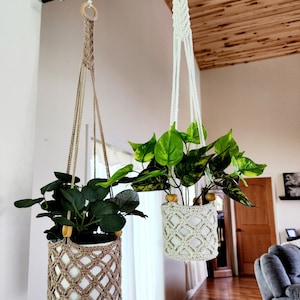 May include: Two macrame plant hangers with wooden rings, each holding a potted plant. The plant hangers are made of natural fiber rope and have a diamond pattern. The plants are green and leafy.
