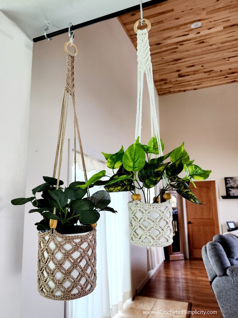 May include: Two macrame plant hangers with wooden rings, each holding a potted plant. The plant hangers are made of natural fiber rope and have a diamond pattern. The plants are green and leafy.