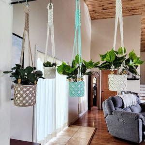 May include: Four macrame plant hangers with different colored pots hanging from a black metal rod. The pots are filled with green plants. The hangers are made of white, beige, and light blue macrame.