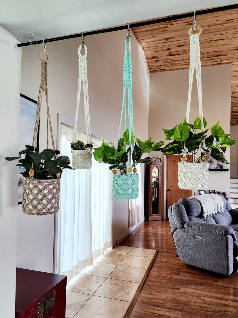 May include: Four macrame plant hangers with different colored pots hanging from a black metal rod. The pots are filled with green plants. The hangers are made of white, beige, and light blue macrame.