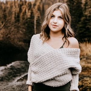 May include: A woman wearing a light gray off-the-shoulder sweater with a textured knit pattern. She is standing in front of a blurred background of trees and a river.