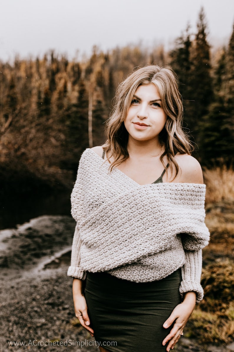 May include: A woman wearing a light gray off-the-shoulder sweater with a textured knit pattern. She is standing in front of a blurred background of trees and a river.