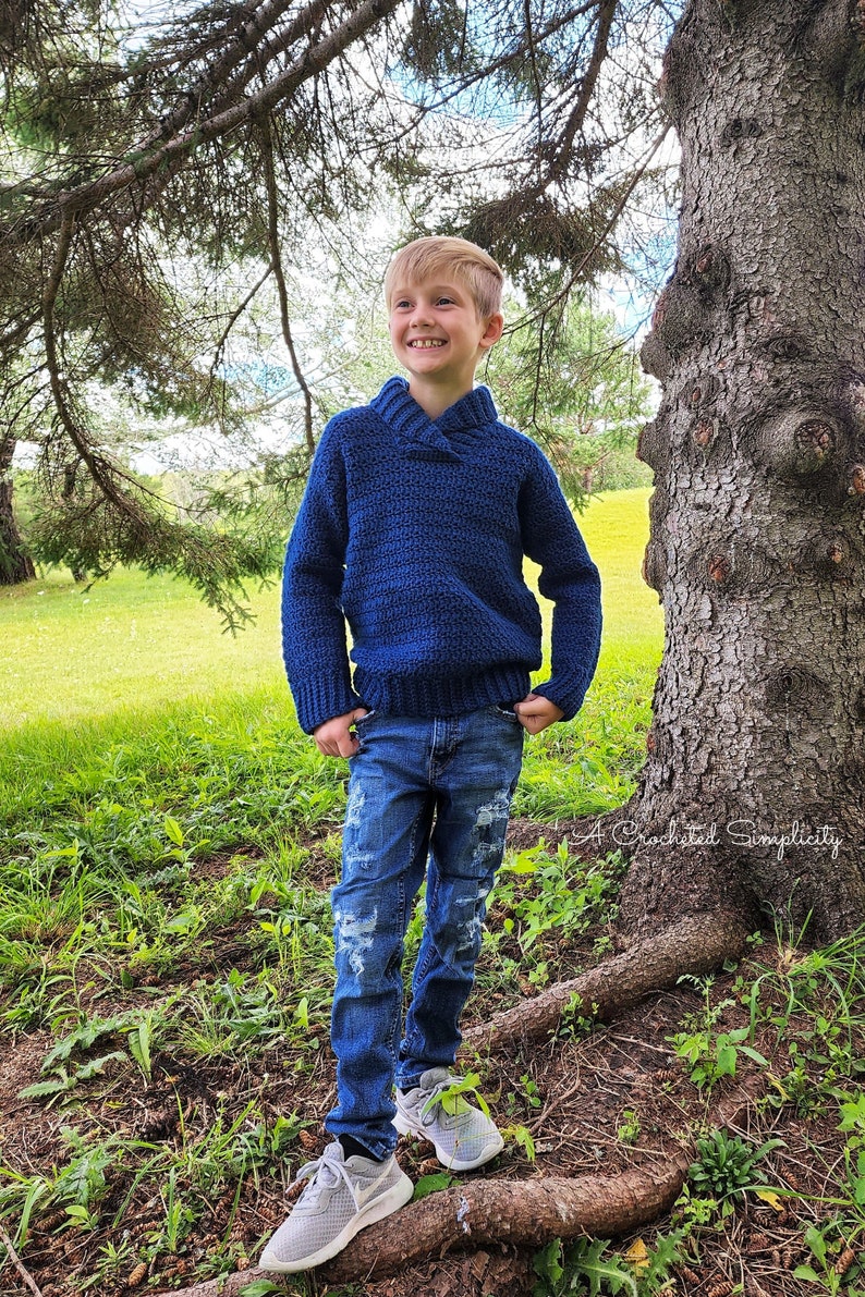 May include: A young person wearing a blue crocheted sweater with a collar, blue jeans, and gray sneakers. The person is standing on a tree root in a grassy area.