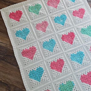 May include: A handmade crochet blanket featuring a repeating pattern of colorful hearts. The blanket is primarily white, with pink, blue, and green hearts arranged in a grid pattern. The yarn appears soft and the stitches are visible.