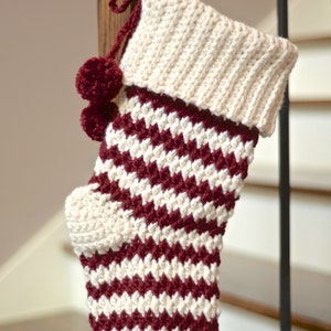 May include: A crocheted Christmas stocking with a white and burgundy chevron pattern. The stocking has a white cuff and a burgundy pom-pom at the top.