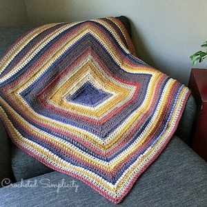 May include: A crocheted blanket with a diamond pattern in shades of blue, yellow, red, and gray. The blanket is draped over a gray armchair. The text "A Crocheted Simplicity" is visible in the lower left corner.