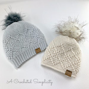 May include: Two crocheted hats with faux fur pom poms. The hats are gray and beige with a textured stitch pattern. Each hat has a brown leather tag with the word "Cozy" on it.