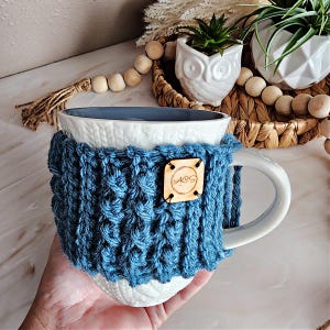 May include: White ceramic mug with a blue knitted cozy. The cozy has a wooden tag with the word "MOS" on it.