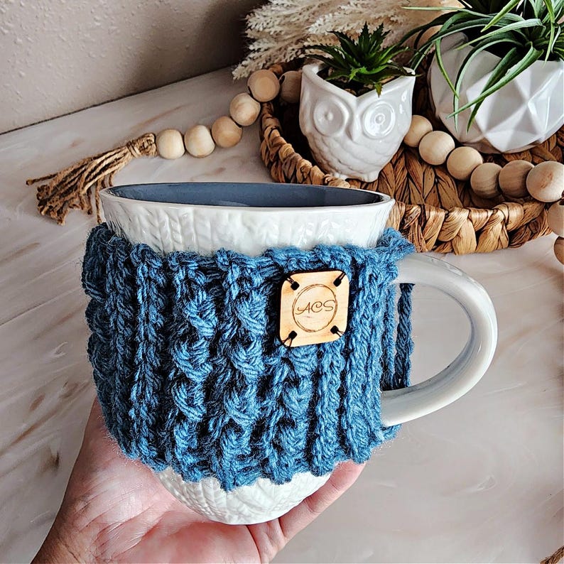 May include: White ceramic mug with a blue knitted cozy. The cozy has a wooden tag with the word "MOS" on it.