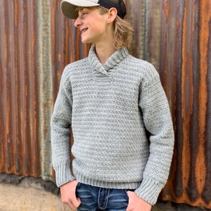 Pu&ograve; includere: Un maglione all'uncinetto grigio chiaro con collo a scialle e polsini a coste. Il maglione &egrave; abbinato a jeans blu e un berretto da camionista grigio e nero. Lo sfondo &egrave; un muro di metallo ondulato.