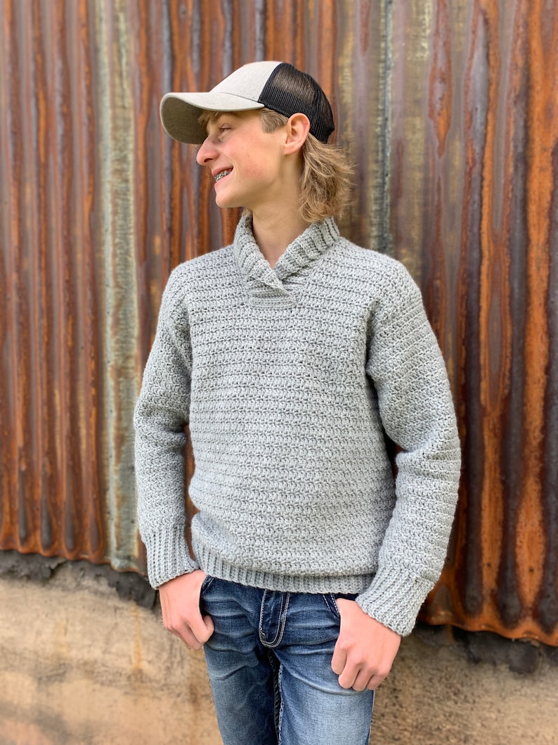 Pu&ograve; includere: Un maglione all'uncinetto grigio chiaro con collo a scialle e polsini a coste. Il maglione &egrave; abbinato a jeans blu e un berretto da camionista grigio e nero. Lo sfondo &egrave; un muro di metallo ondulato.