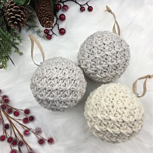 May include: Three knitted Christmas baubles in shades of grey and cream, each with a hanging loop. The baubles are arranged on a white, fluffy surface, with pine cones and red berries in the background.
