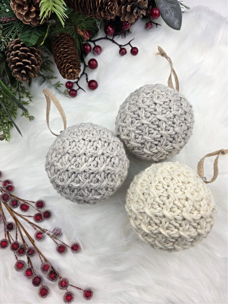 May include: Three knitted Christmas baubles in shades of grey and cream, each with a hanging loop. The baubles are arranged on a white, fluffy surface, with pine cones and red berries in the background.