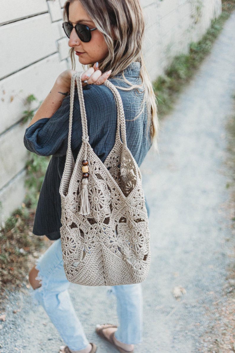 May include: A beige crochet tote bag with a floral design. The bag has long straps and a tassel with wooden beads. The bag is carried over the shoulder. The bag is made of a soft, woven material.