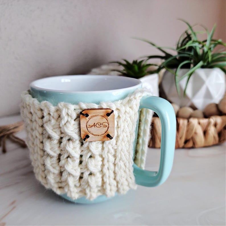 May include: A light blue ceramic mug with a white crocheted cozy. The cozy has a wooden tag with the text "ACS" on it.