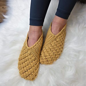 Op de afbeelding: Een paar handgemaakte, mosterdgele gehaakte pantoffels. De pantoffels hebben een textuur met ribbels en een comfortabel ontwerp met open bovenkant. De pantoffels worden getoond op een witte, pluizige ondergrond, met donkerblauwe leggings op de achtergrond.