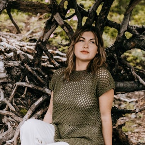 May include: A woman wearing a green crochet top with a white crochet design. She is sitting in a natural setting with branches and twigs surrounding her.