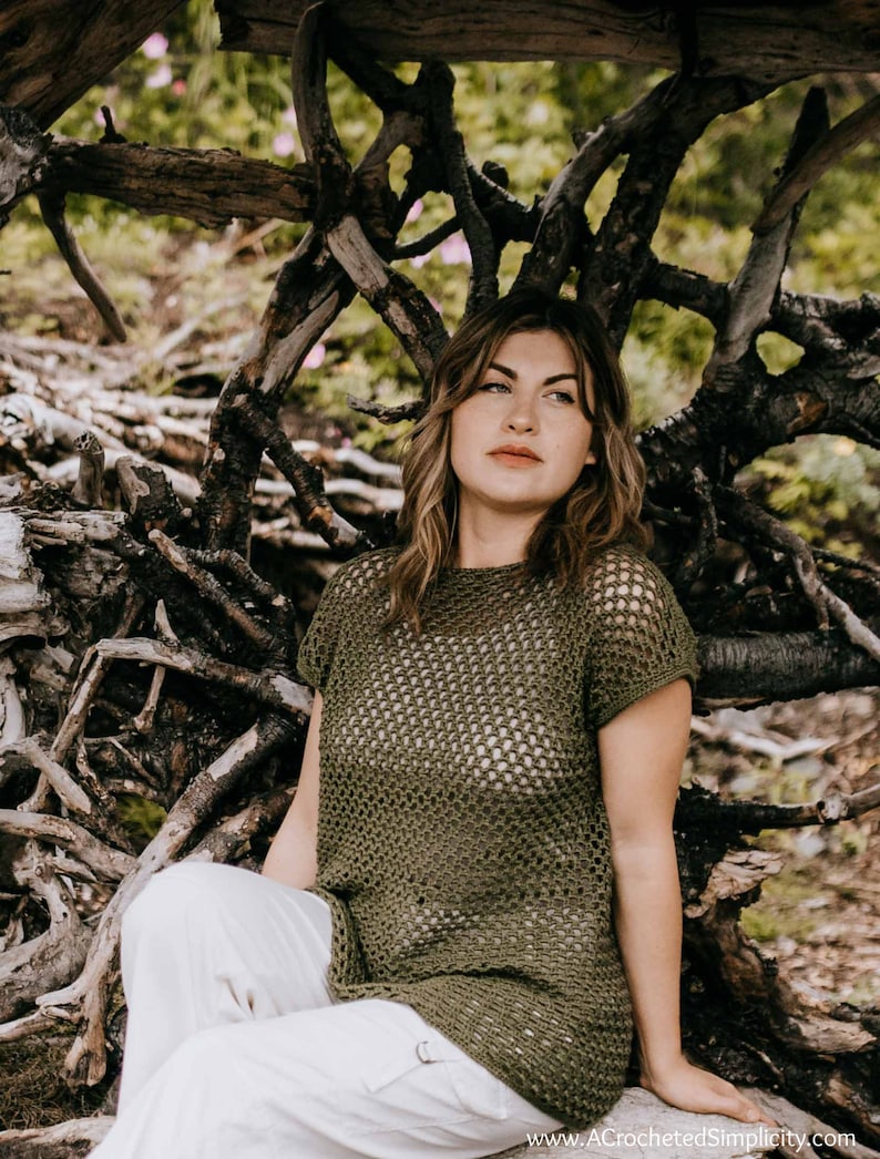 May include: A woman wearing a green crochet top with a white crochet design. She is sitting in a natural setting with branches and twigs surrounding her.